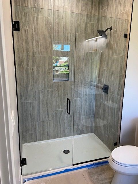 Shower stall with glass doors, black hardware, gray tile, and a white base, next to a toilet.