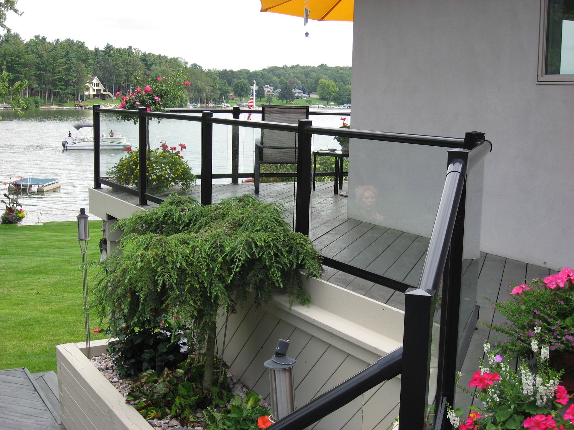 Deck with glass railing overlooks a lake with boats. Black railing, yellow umbrella, and greenery.