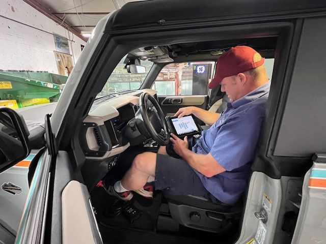 A man in a maroon cap uses a tablet inside a light gray Ford Bronco.