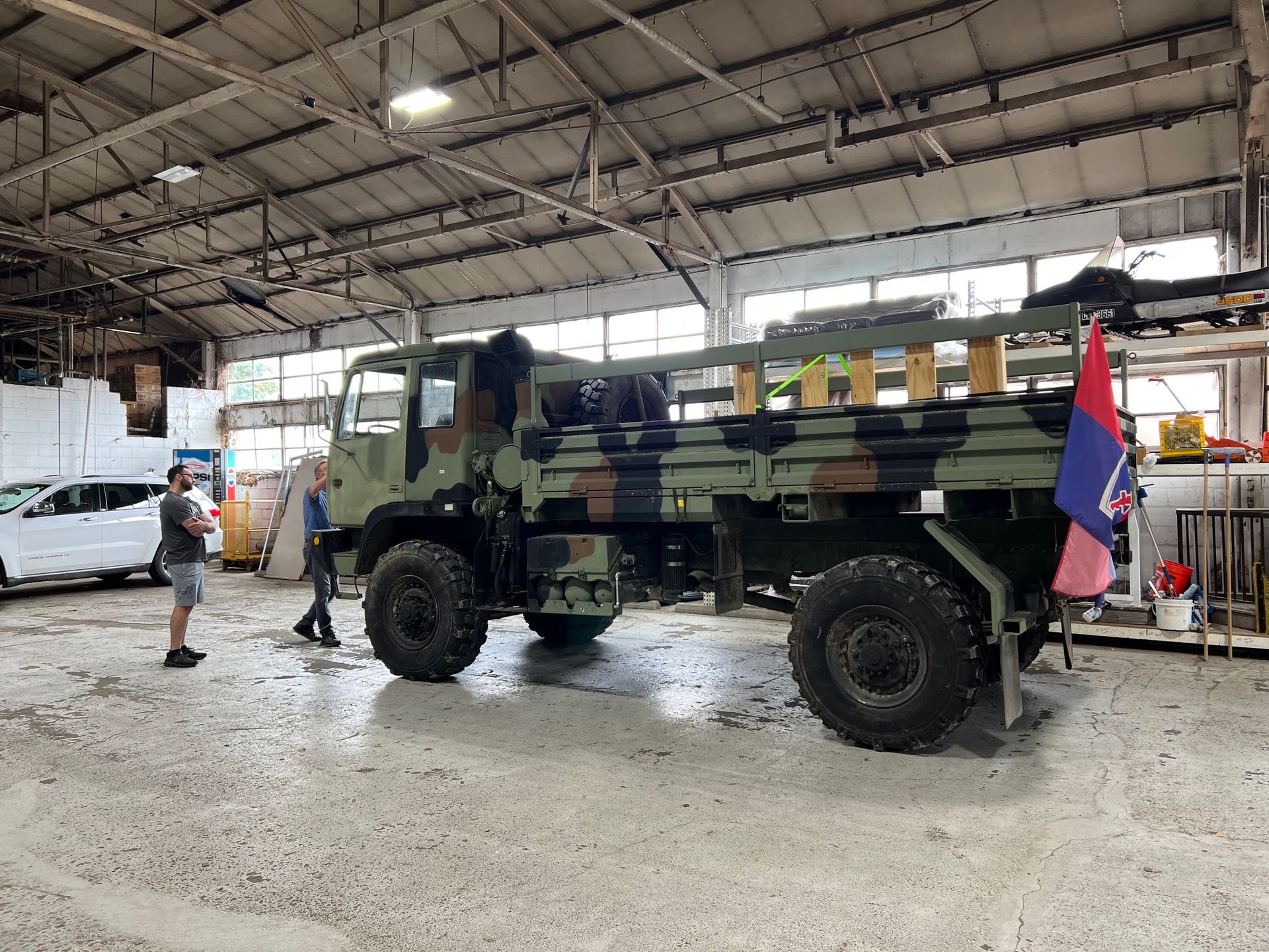 Camouflaged military truck inside a large building; two people stand nearby.