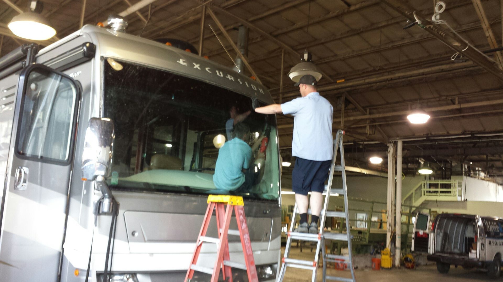 Two people replacing a windshield on a large RV in a garage. One on a ladder reaching up.
