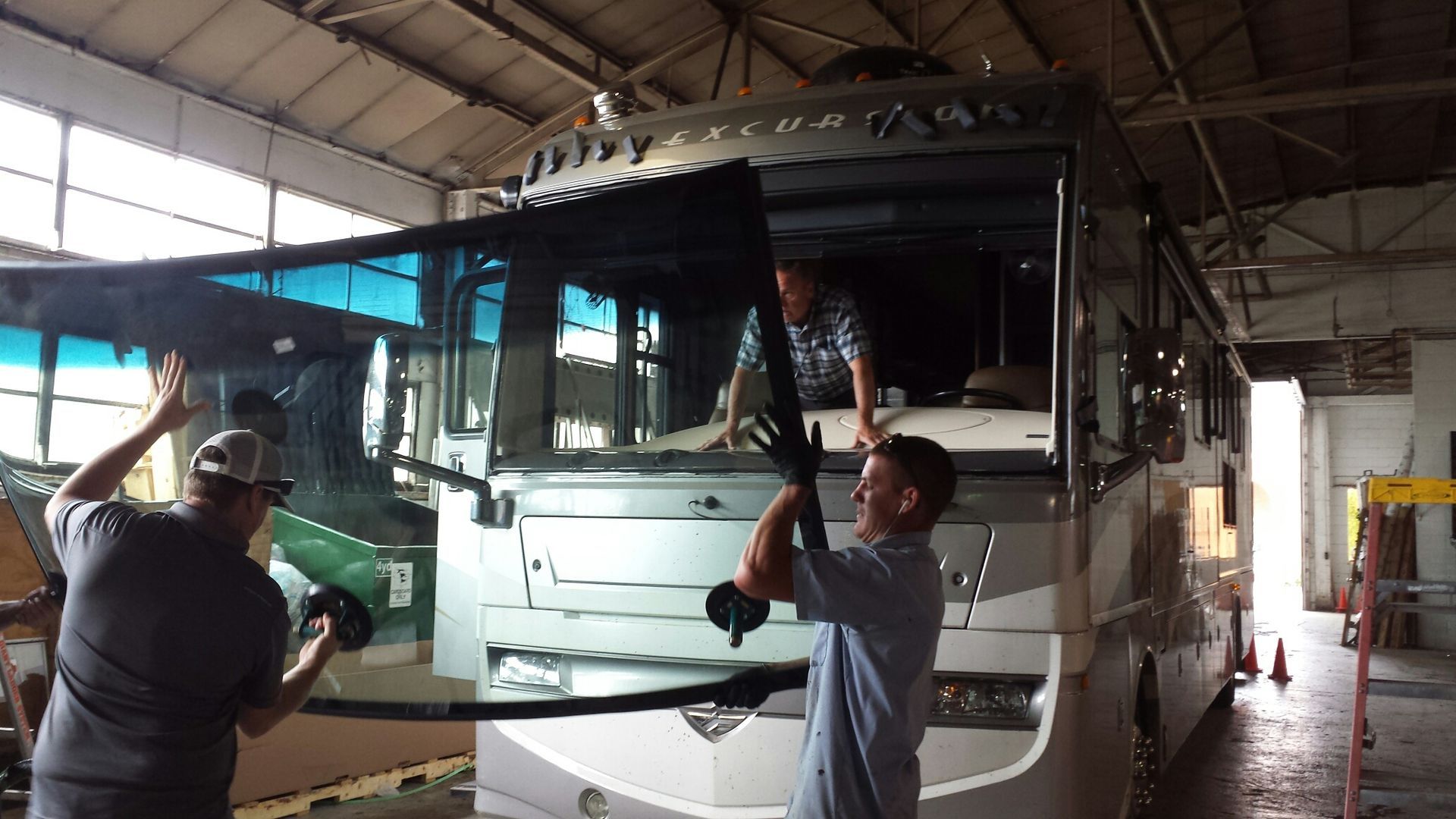 Three workers removing a windshield from a large silver RV inside a garage.