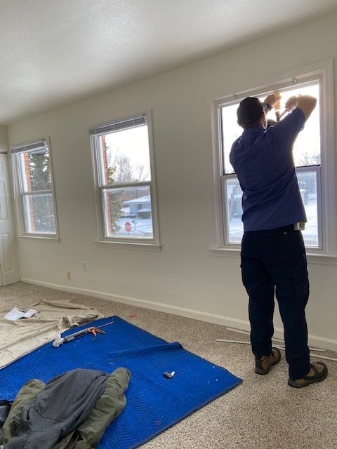 Man installing a window in a room with three windows. Blue tarp and tools on the floor.