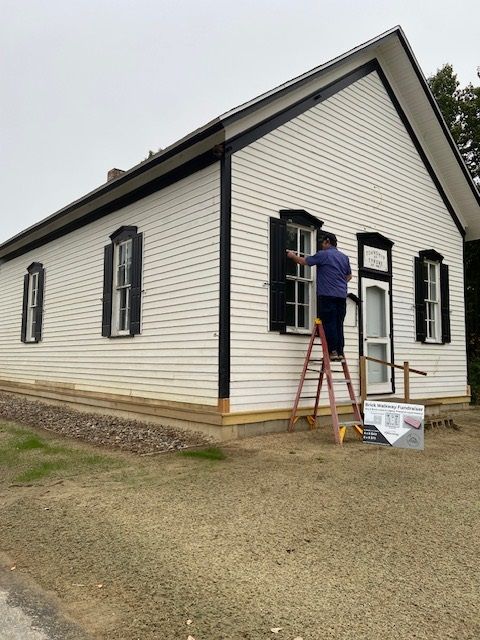 Person on ladder adjusting black shutter on white building with black trim. Outdoors.