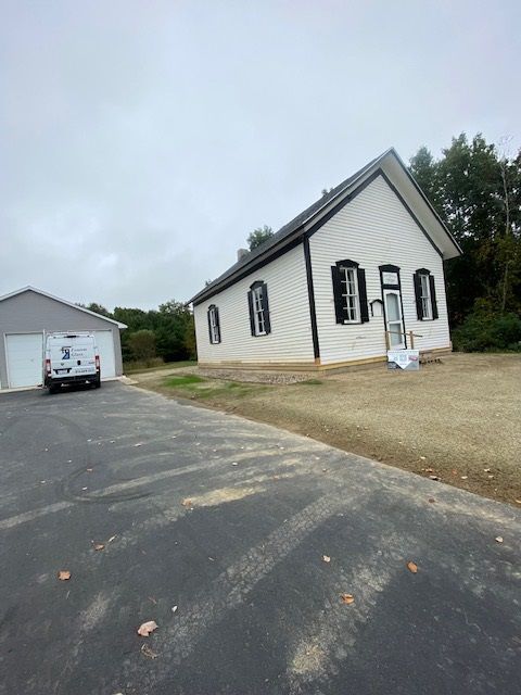 White church-like building with black trim and a garage on a driveway under an overcast sky.