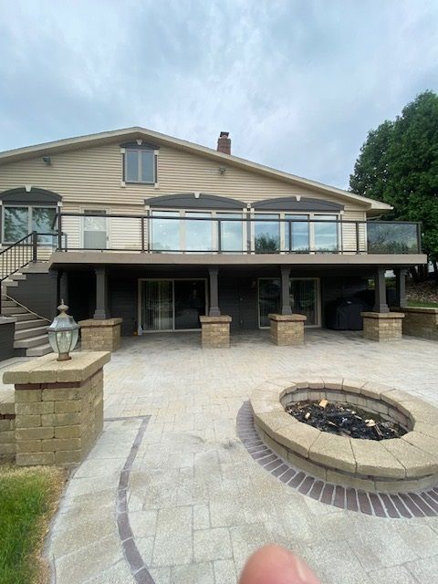 Back of a tan house with a dark deck and patio, a fire pit, and glass railing on deck.