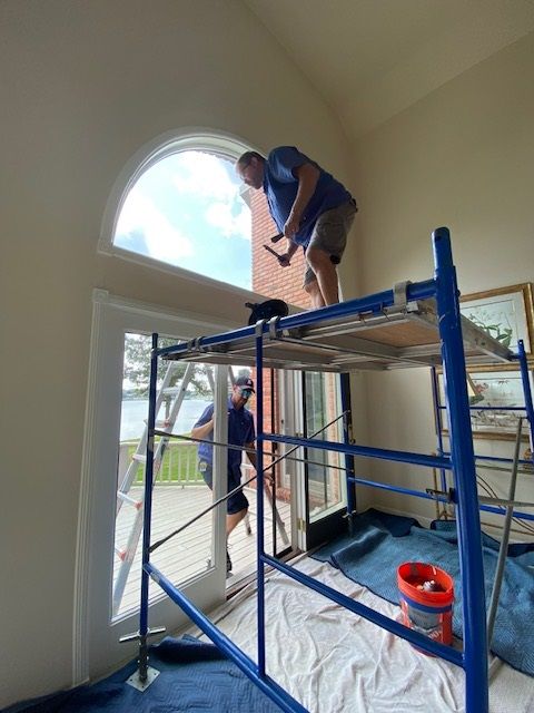Man on scaffolding repairs window, another man stands below, inside a house.