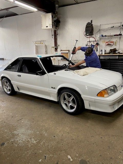 A white Ford Mustang being worked on by a person in a shop setting, replacing windshield.