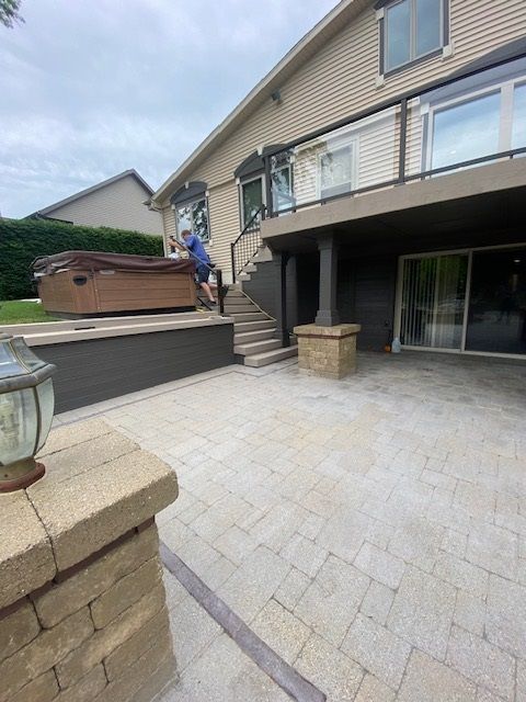 Person climbing stairs to a raised patio with hot tub, beige brick pavers, and a two-story house.
