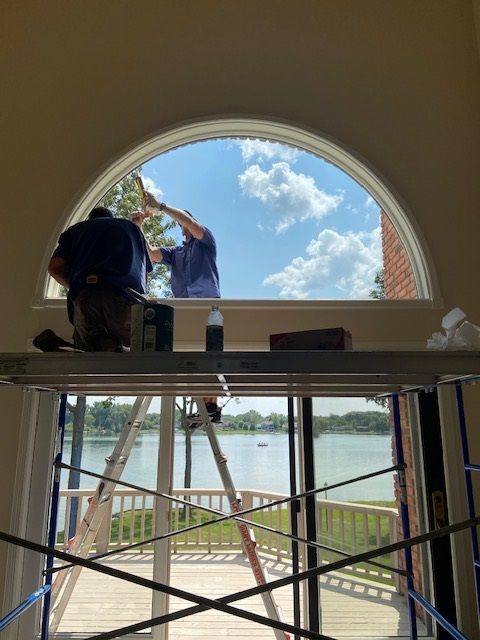 Two men cleaning a large arched window. One on a scaffold, the other inside. Sunny day, water view.
