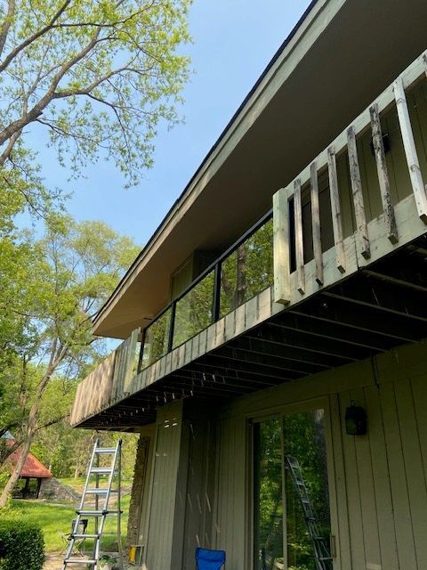 A two-story house with a wooden deck and glass railing. A ladder leans against the siding.