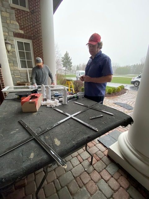 Two men repairing window panes on a table, under a porch with columns. Overcast day.