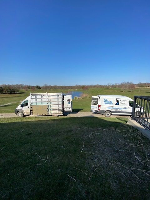 Two white work vans parked on grass, one with a glass rack. Clear blue sky.
