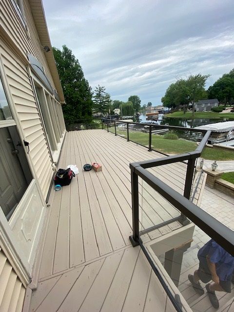 Deck with glass railings overlooking a body of water. Beige siding on the house. Overcast sky.