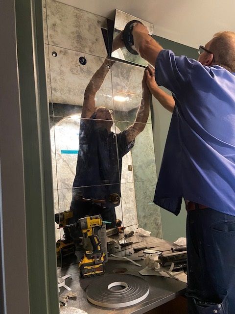 Two men installing a large mirror. One holds the top edge; the other works overhead. Tools sit on the counter.