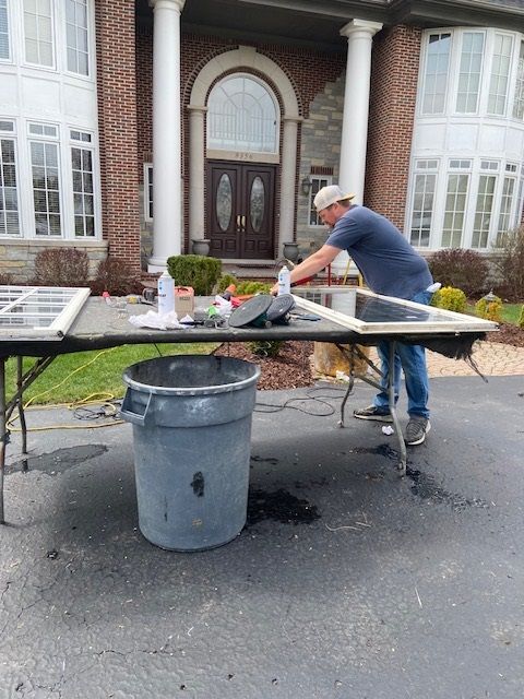 Man working on window frames outdoors in front of a brick house. A trash can and table are nearby.