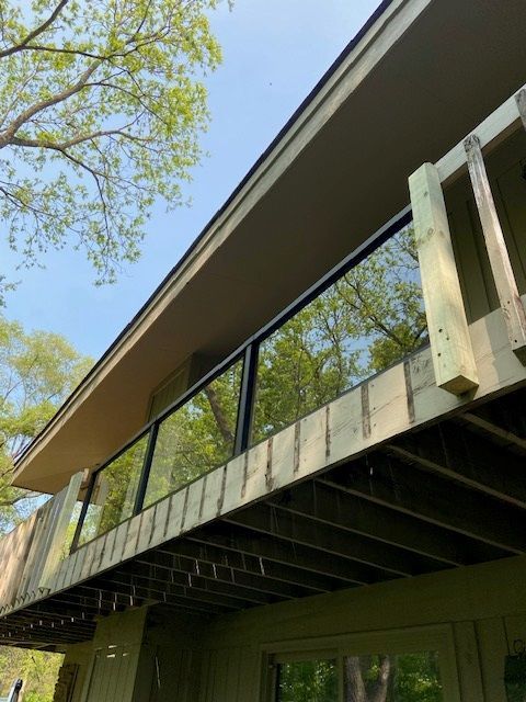 Deck with glass railing, overlooking trees. Beige roof overhang, natural wood deck, and blue sky.