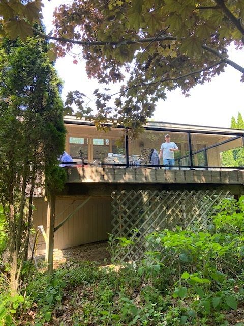 Person standing on a deck, surrounded by trees. The house has large windows and a lattice base.