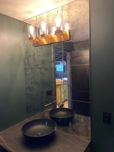 Bathroom with two black vessel sinks, large mirror, gold light fixture, and dark green walls.