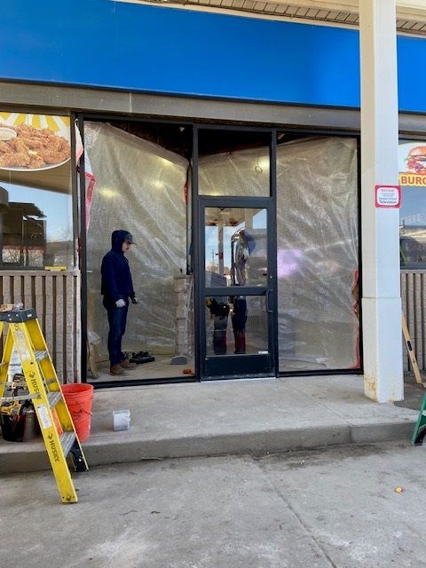 Construction at a storefront; people inside, wrapped in plastic. A ladder, bucket, and tools are visible.