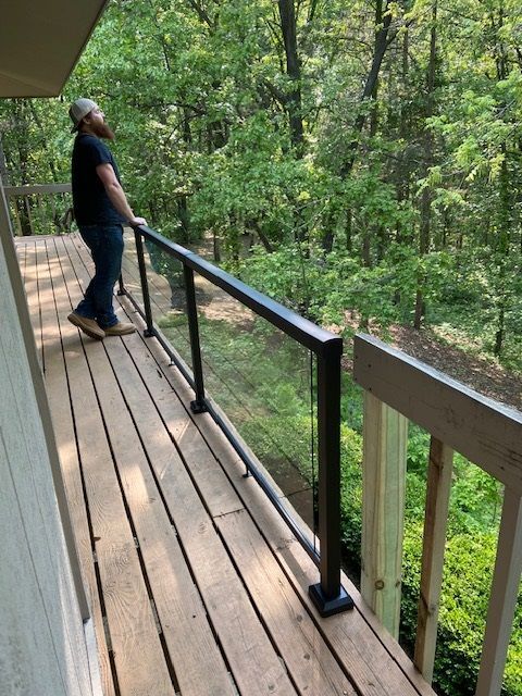 Man on a wooden deck with glass railing, looking up at trees in a forest.
