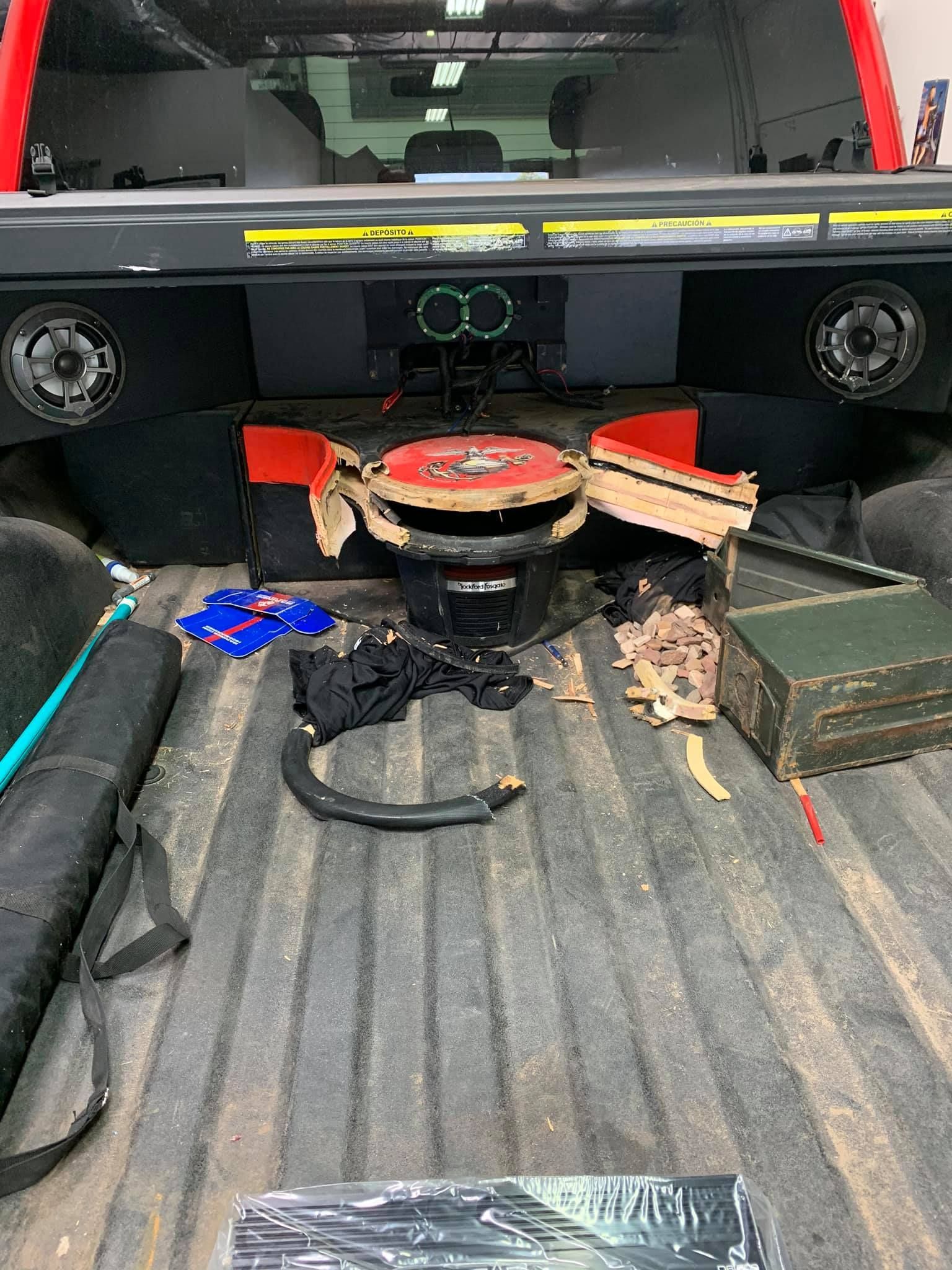 A truck bed with a large subwoofer and speakers installed. Cardboard and other items are strewn about.