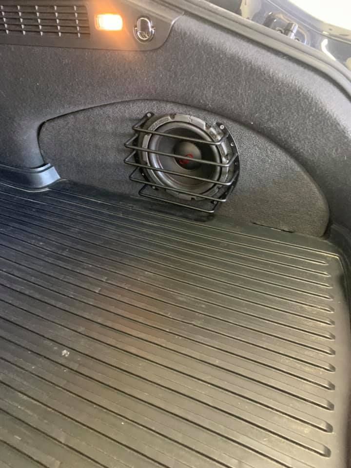 A car's trunk with a subwoofer speaker protected by a metal cage mounted on the side. A rubber mat lines the floor.