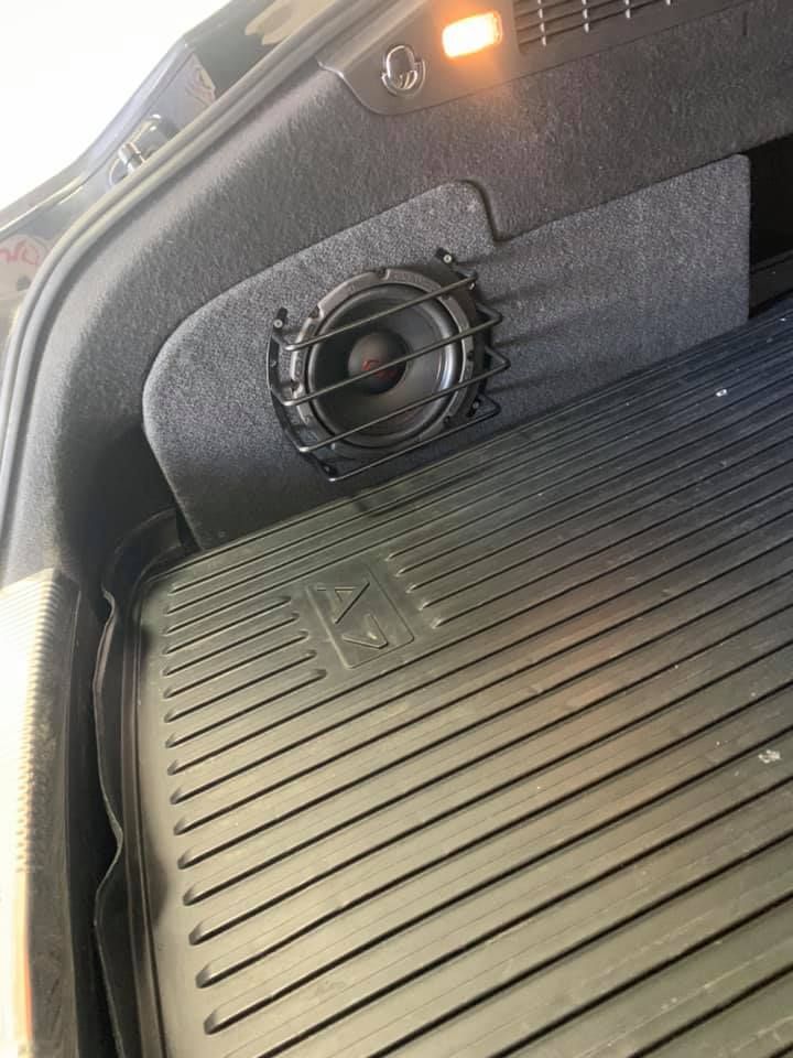 Black speaker in the trunk of a car, mounted on a gray fabric panel. A black rubber mat is below.