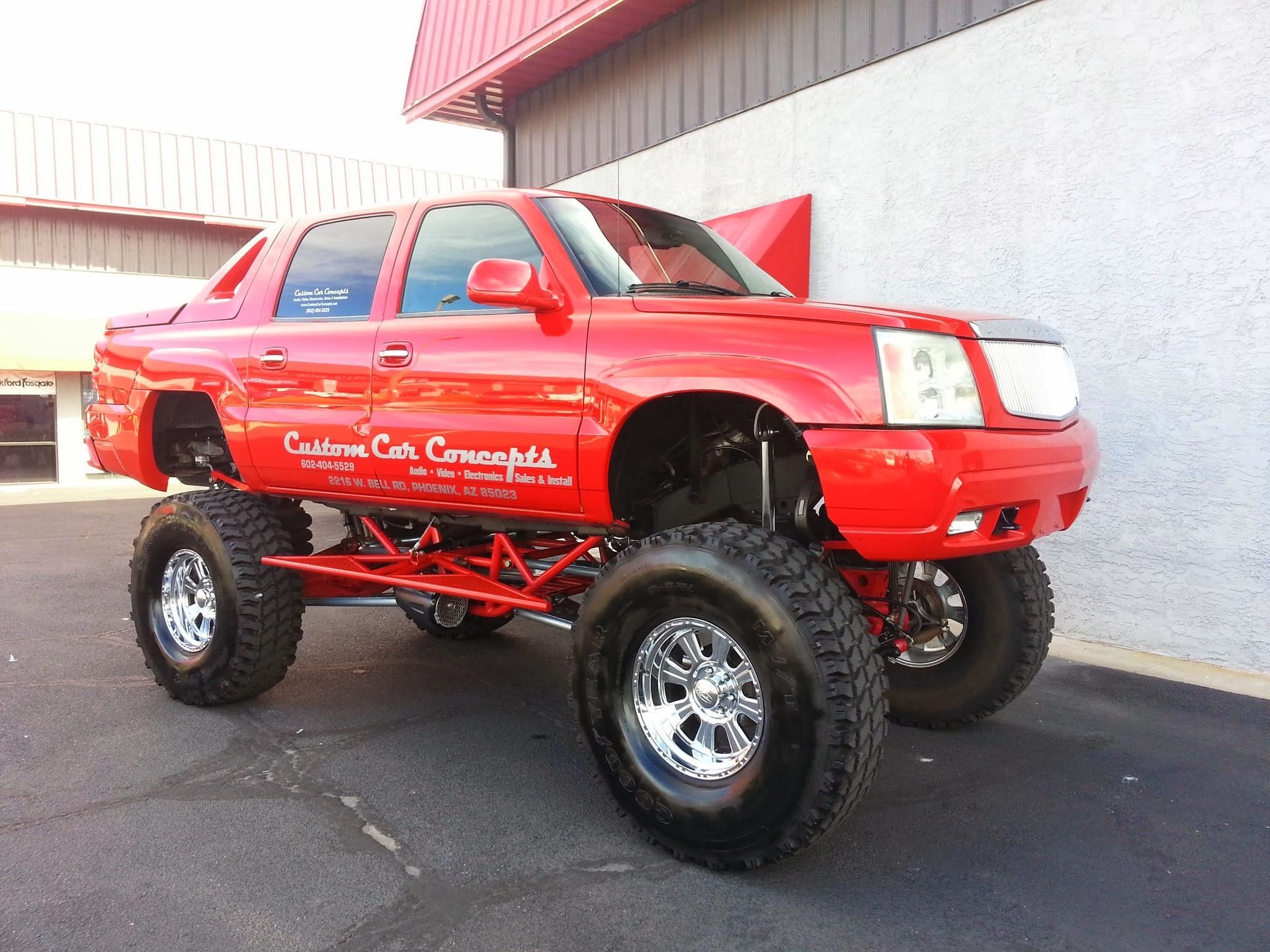 Red monster truck, a lifted Cadillac Escalade, parked in front of a building. It has large tires and chrome wheels.
