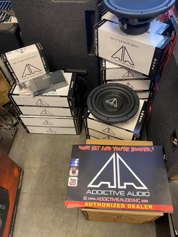 Stack of car audio subwoofers and amplifiers in boxes from Addictive Audio, displayed in an auto shop.