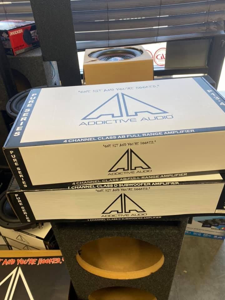 Two boxes of Addictive Audio car amplifiers stacked on a speaker box inside a store.