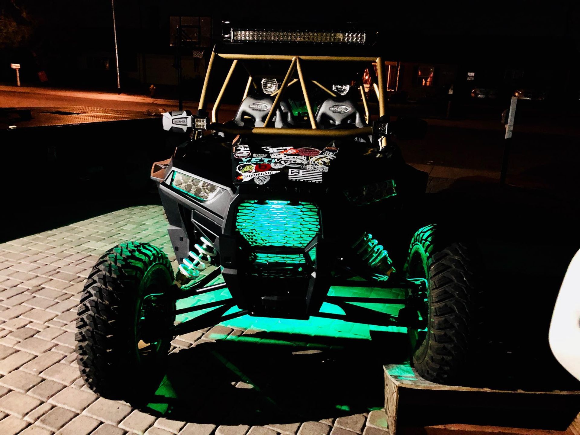 Black off-road vehicle with a roll cage and green underglow, parked on a brick surface at night.