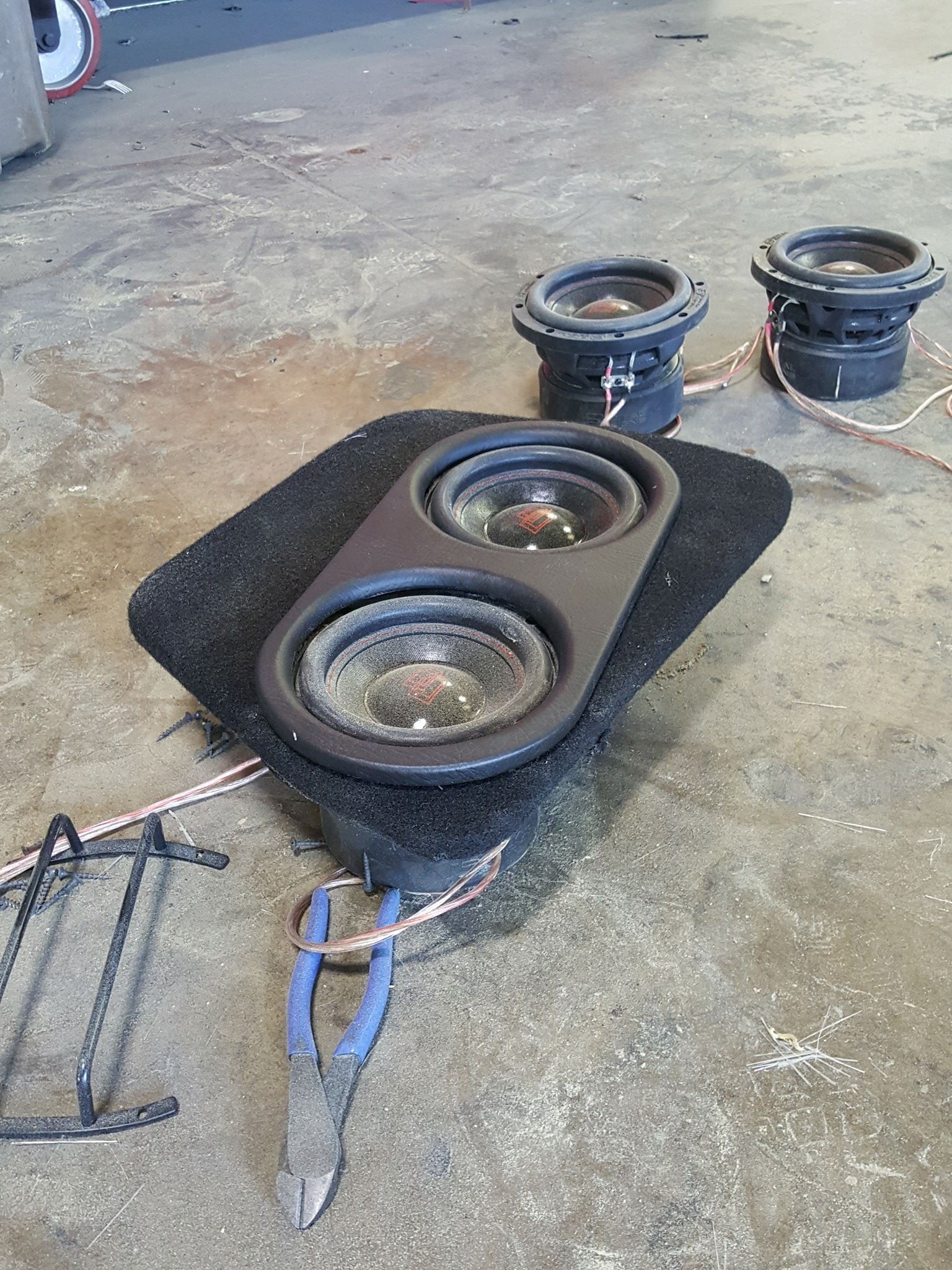 Two car subwoofers mounted on a black platform, with two more nearby, connected by copper wiring.