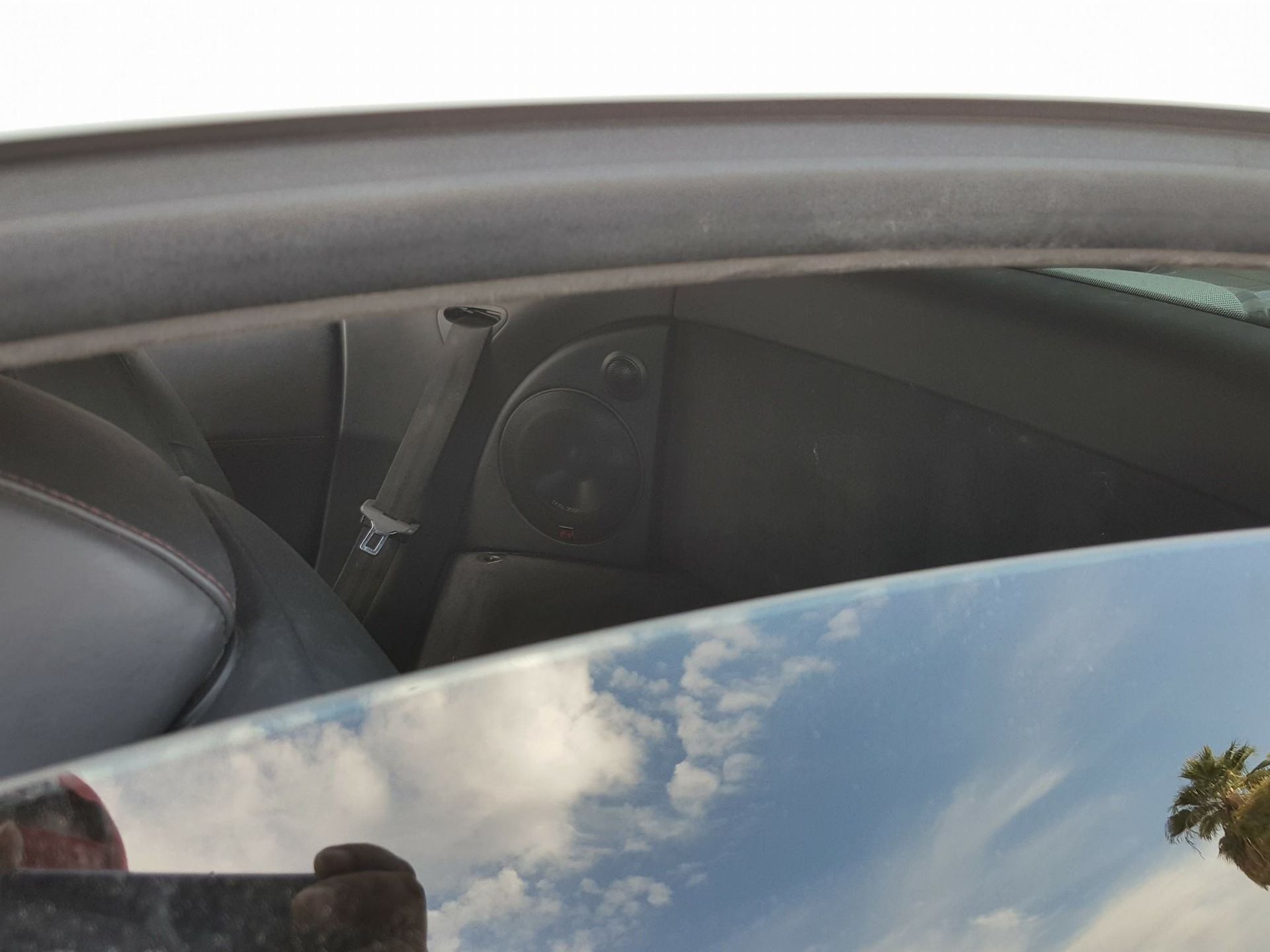 Black car interior with a speaker, seatbelt, and window reflecting clouds.