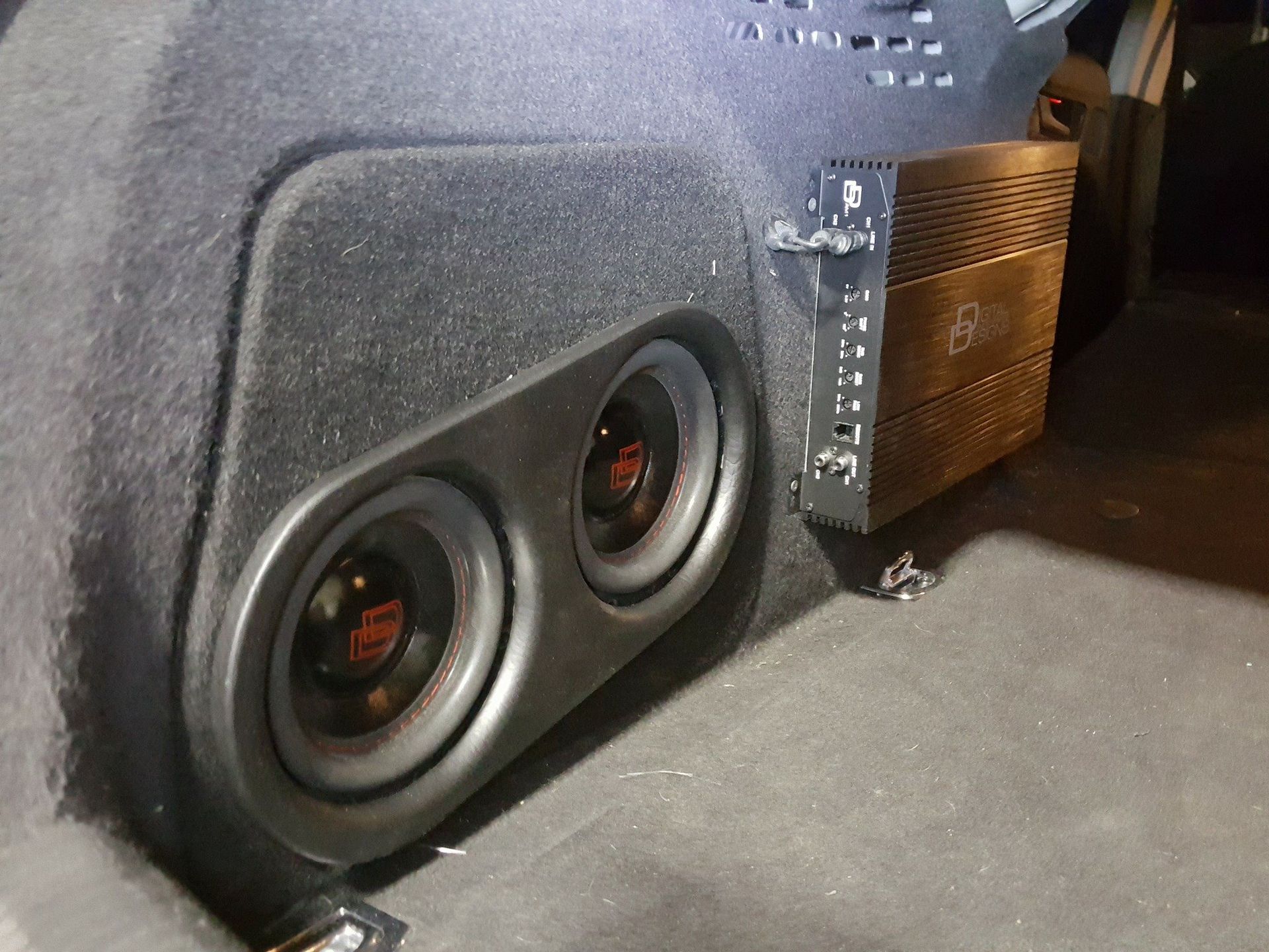 Two subwoofers and an amplifier mounted in the trunk of a car. Black enclosure with dark gray carpet.