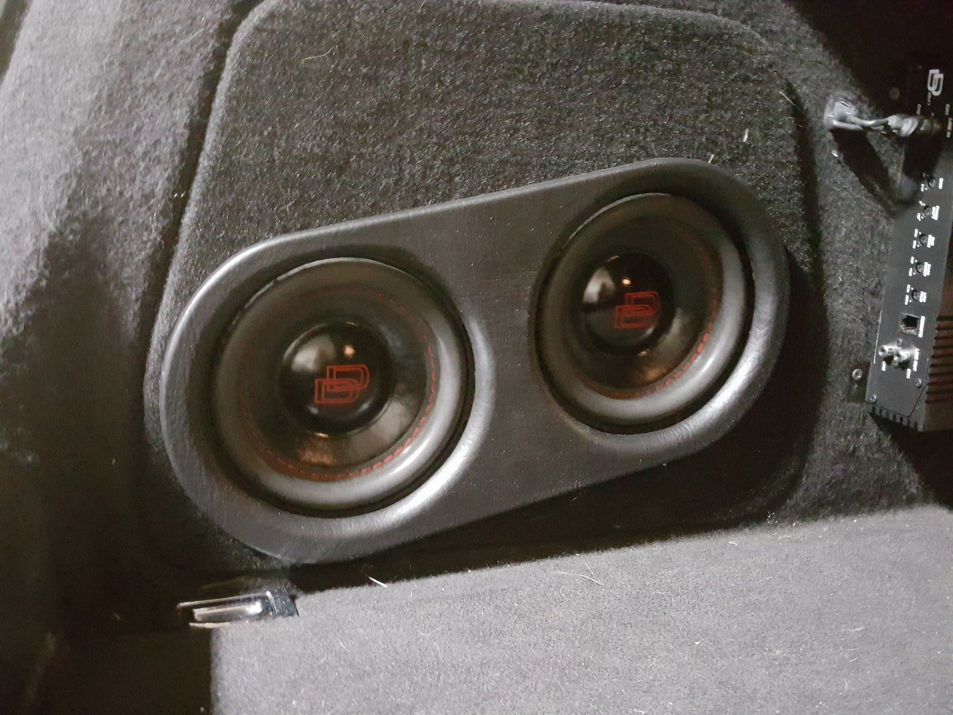 Two black subwoofer speakers mounted in a vehicle's trunk against a dark gray fabric panel.