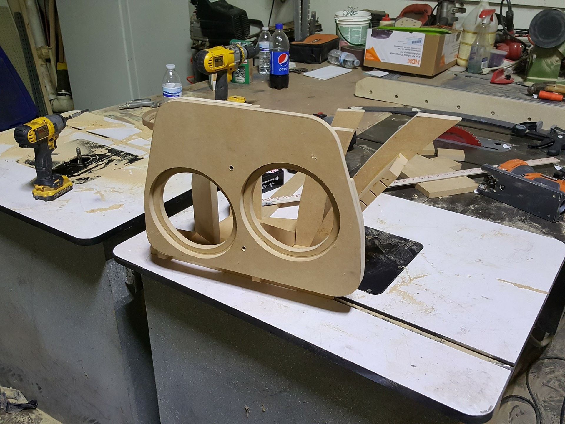 A partially built wooden speaker enclosure with two circular cutouts, resting on a workbench in a cluttered workshop.