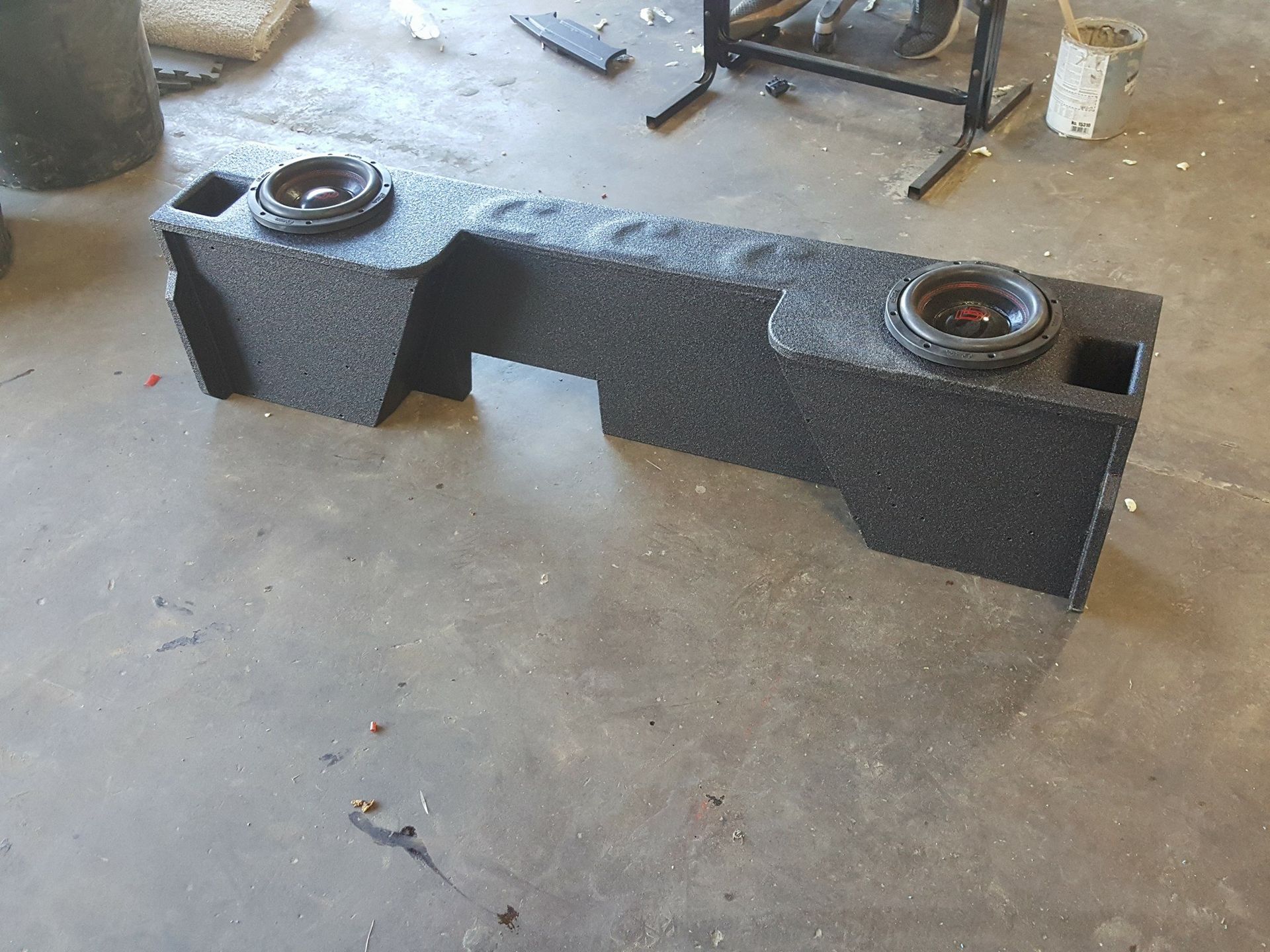 A custom black subwoofer box with two speakers built for a vehicle, resting on a concrete floor.