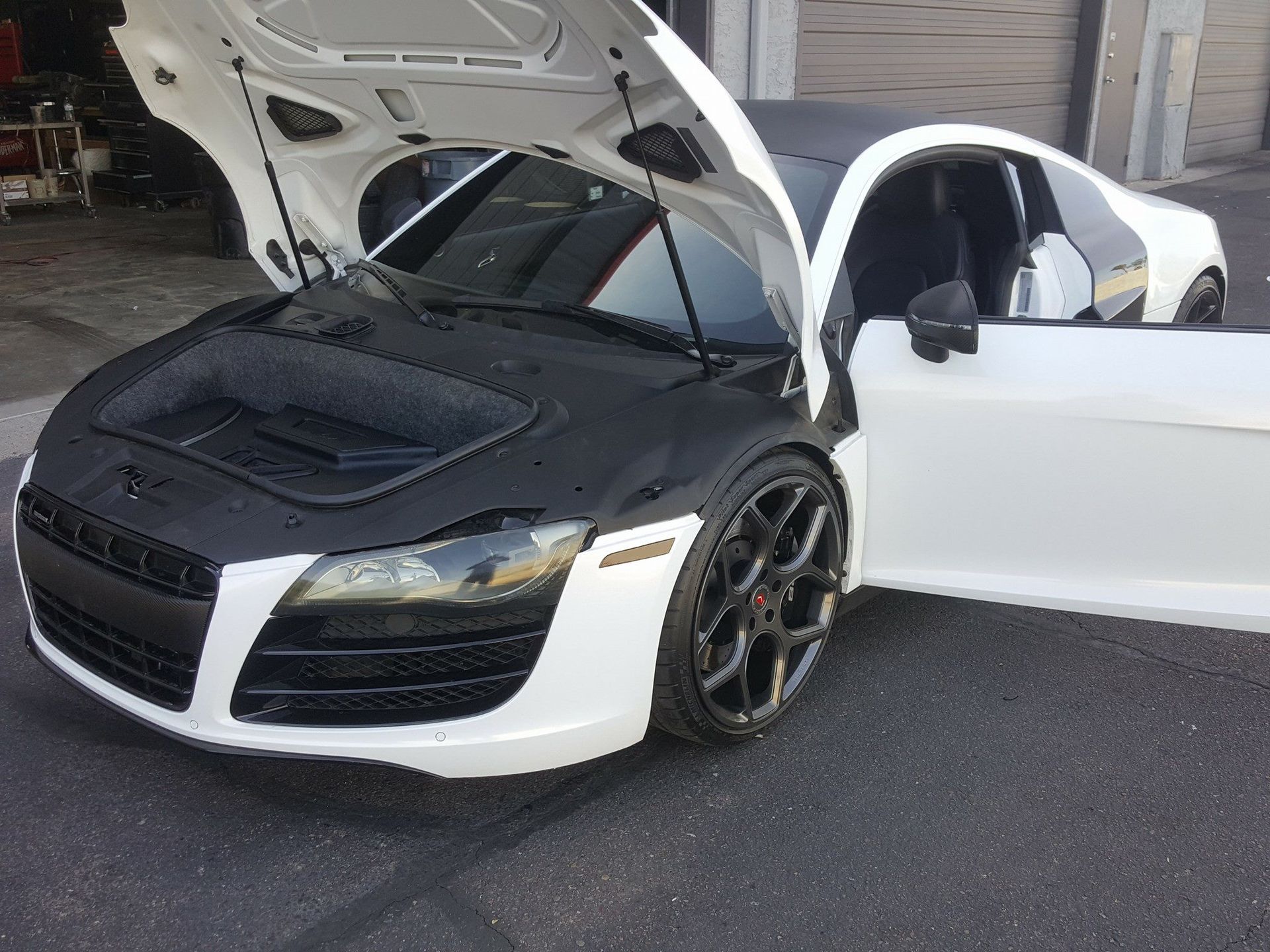 White Audi R8 sports car with a black hood, and black wheels, parked with the hood and door open.