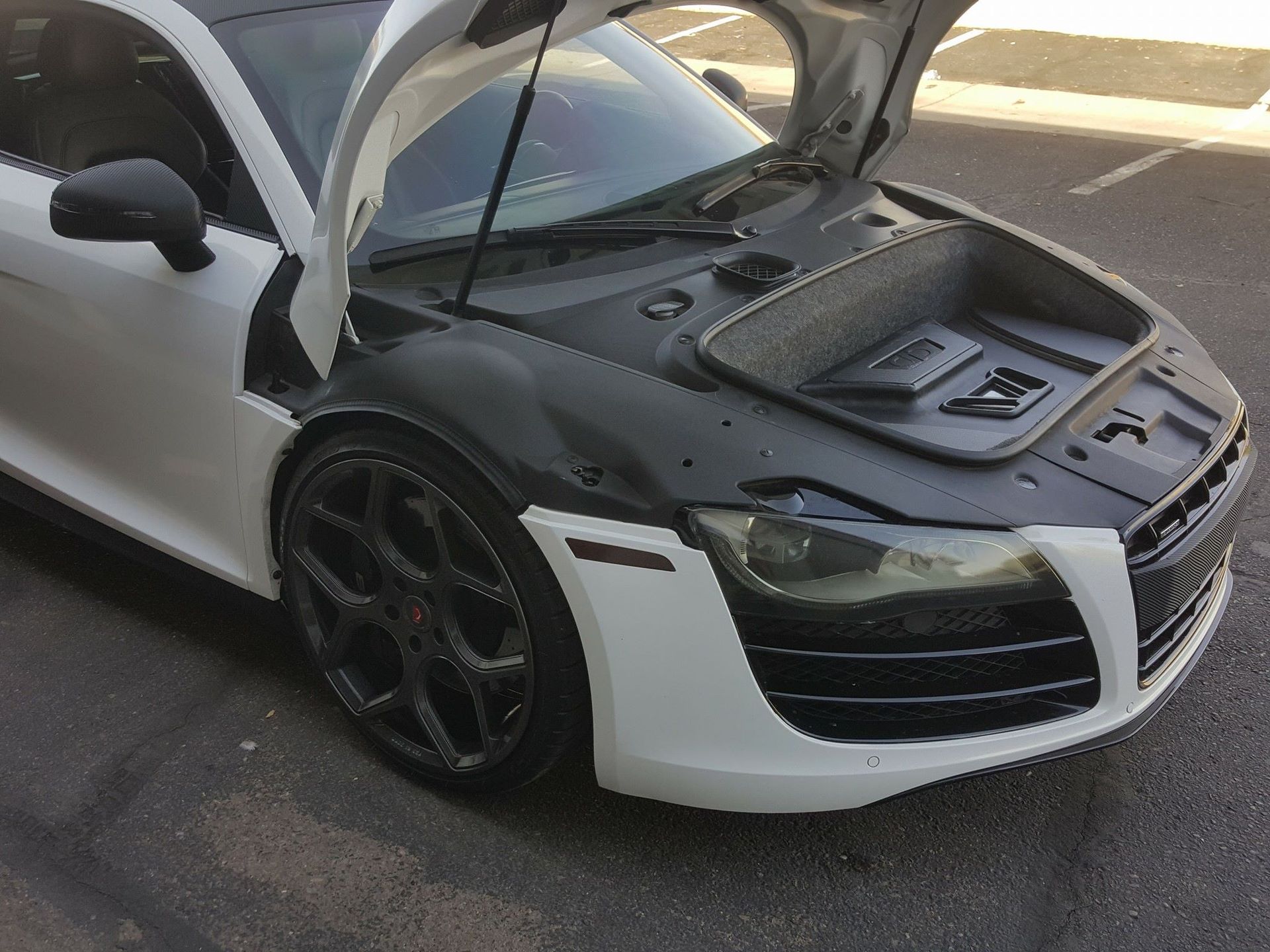 White Audi R8 with the hood open, revealing a black interior. Black wheels and side details are visible.