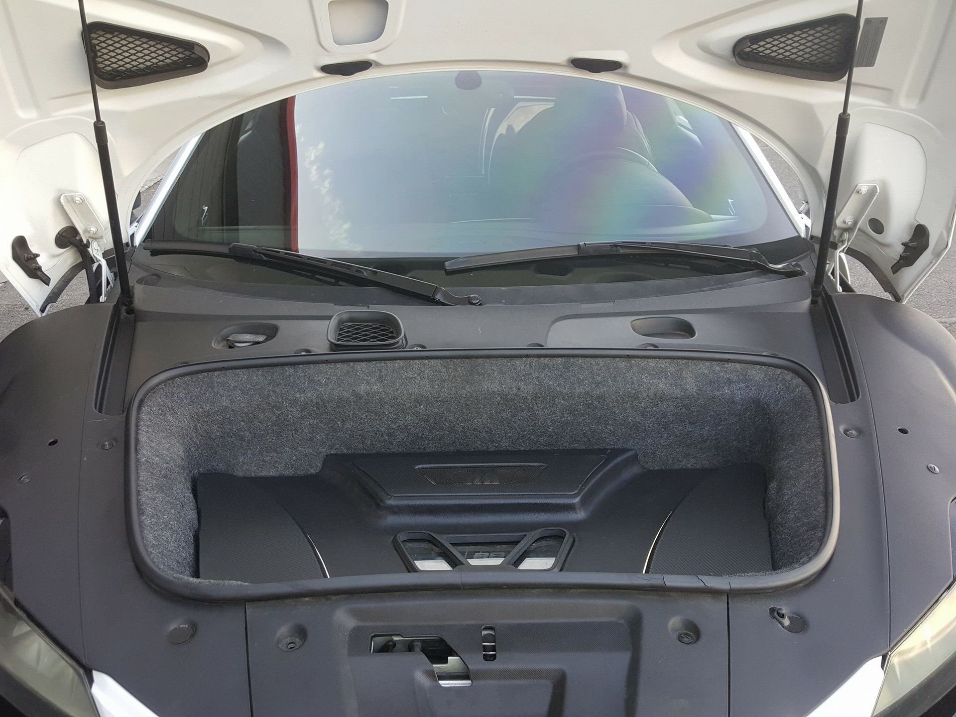 Open front trunk of a white sports car, revealing a gray lined compartment. The car is outside on a sunny day.