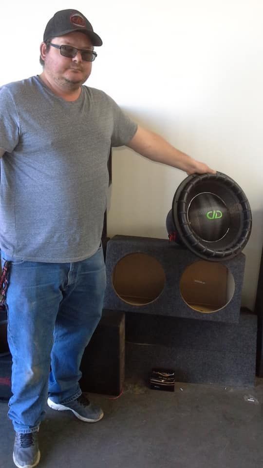 Man in a gray shirt and jeans stands next to a double subwoofer box and a speaker. Indoors, garage-like setting.