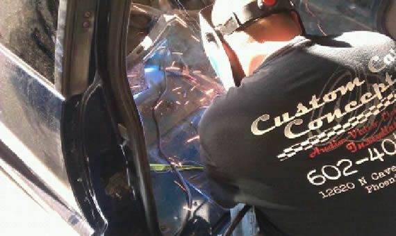 Person welding a car door; sparks flying. They wear a welding helmet and a black shirt with 