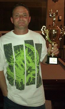 Man in a white t-shirt holds a gold trophy. He is indoors and smiles slightly.