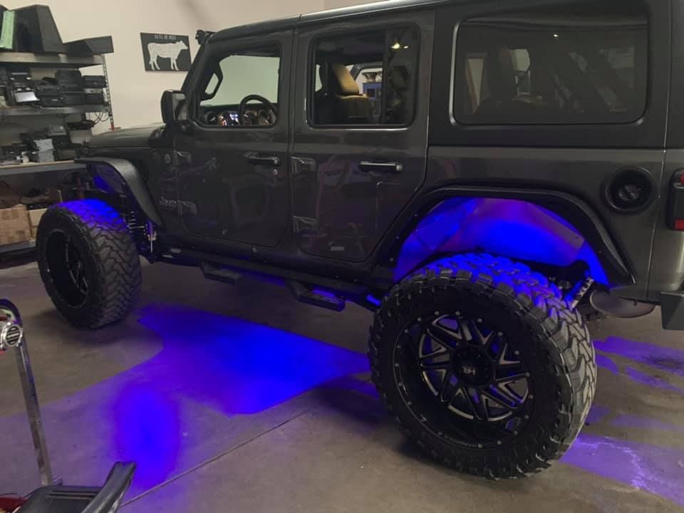 Dark gray Jeep Wrangler with large black wheels and blue underglow lights. It's parked inside a garage.