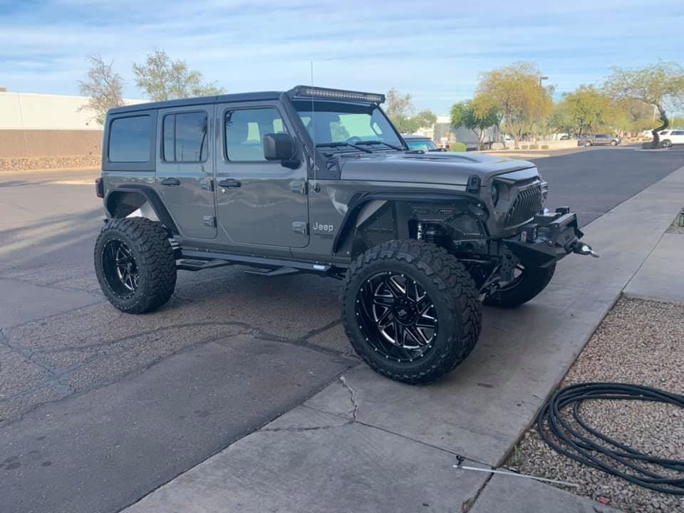 A gray Jeep Wrangler modified with large black wheels and tires, a light bar, and a custom bumper, parked on a paved street.