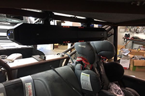 Black speaker mounted inside a vehicle with a child car seat. Red lights are visible on the speaker.