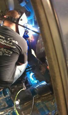A person welds inside a vehicle, using a welding mask and protective gear, with bright sparks visible.
