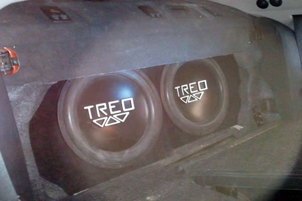 Two Treo subwoofers mounted in a black car trunk. The speakers have white lettering and are centered.