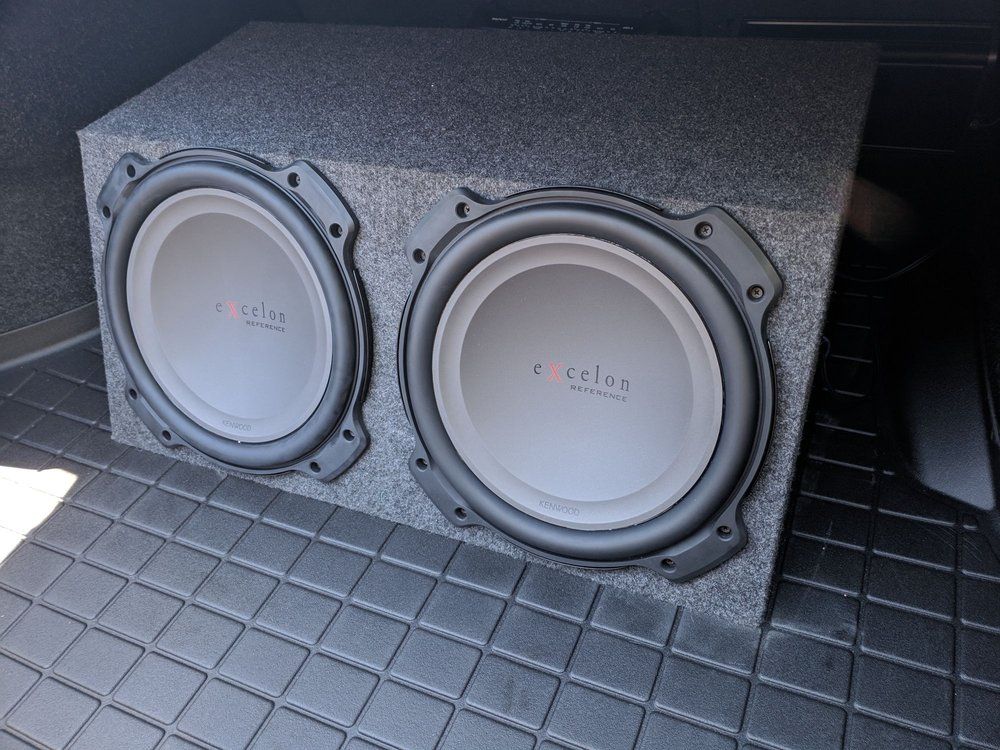 Two white car subwoofers are installed in a gray box, placed in the trunk of a car with a black mat.